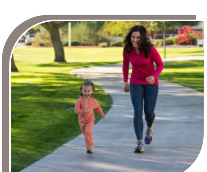 A woman and a young child jogging together on a sidewalk in a sunny park.